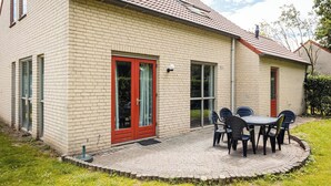 A two-story building with a brick exterior, a red door, and a patio with a table and chairs. - Quiet Woodland Getaway for Families (Ewijk)