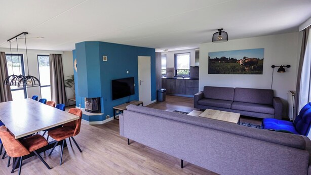 A modern living room with a large grey sofa, a dining table with chairs, a flat-screen TV, and a kitchen area in the background. - Quiet Woodland Getaway for Families (Ewijk)