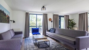 A modern living room with a sofa, a blue chair, a coffee table, and a large window with curtains. - Quiet Woodland Getaway for Families (Ewijk)