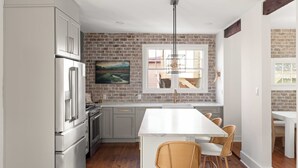 A modern kitchen with grey cabinets, a brick wall, a large window, and a dining area with wicker chairs. - Luxe 5BR w/ Heated Pool~1 Block to King~Lowcountry House (Charleston)