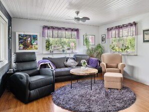 A living room with a black leather sofa, a beige armchair, and a grey couch. There is a round coffee table with a vase of flowers. The room has large windows with purple curtains, a ceiling fan, and a wooden floor. - 4 Star Holiday Home in Bergshamra (Bergshamra)