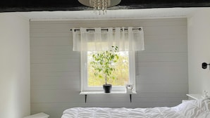 A bedroom with a bed, a small white nightstand, a window with a potted plant, and a white curtain. - 4 Star Holiday Home in Bergshamra (Bergshamra)