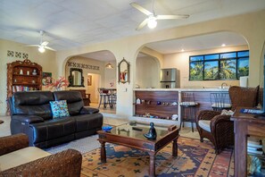 A spacious living area with a black leather sofa, a coffee table, and a dining area with chairs. There is a kitchen with a refrigerator and a window with a view of palm trees. - GARDEN – 2BR Treetop Home Steps from Los Ayala Beach (Los Ayala)