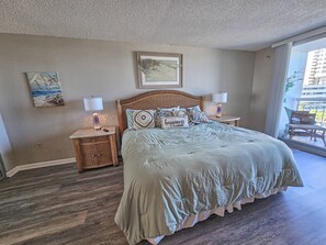 Een slaapkamer met een groot bed, twee nachtkastjes, een rieten hoofdbord, een ingelijst schilderij aan de muur en uitzicht op een balkon.