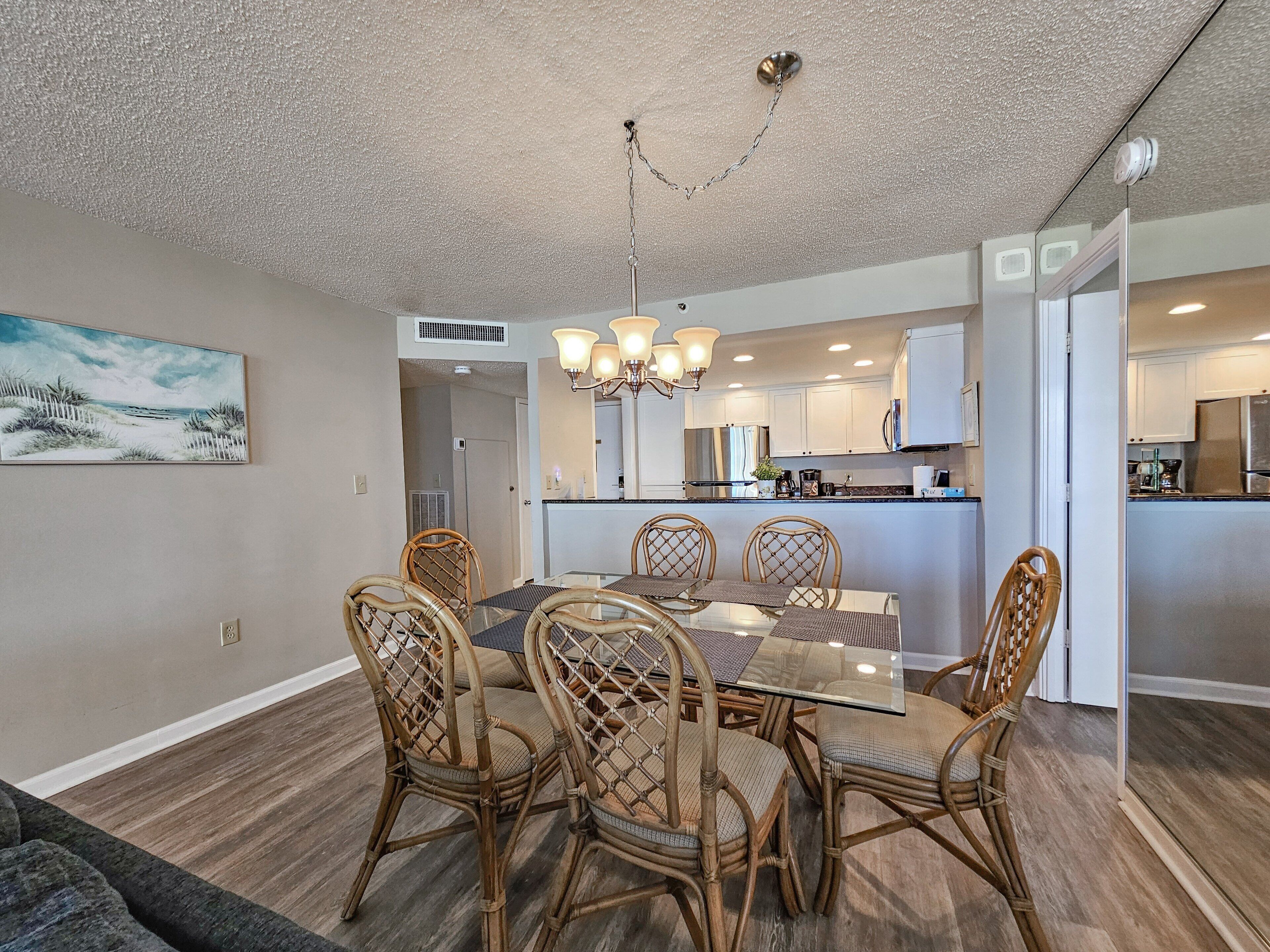 A dining area with a glass table and wicker chairs, a kitchen with white cabinets, and a beach scene painting on the wall.