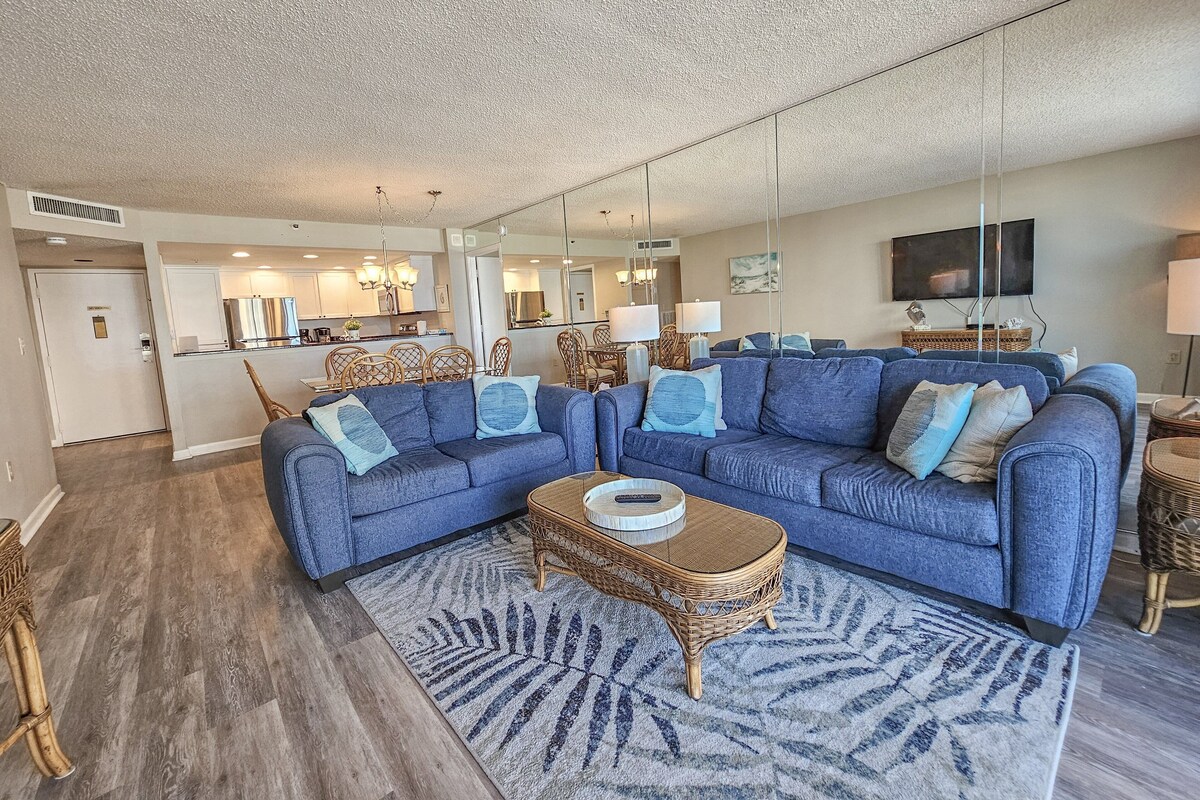 A spacious living room with a large sectional sofa, a coffee table, and a dining area in the background.