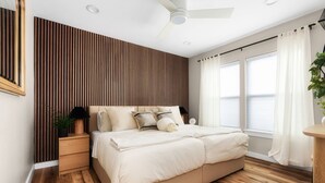 A bedroom with a bed, wooden panel wall, ceiling fan, windows with curtains, and a bedside plant.