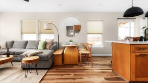 A modern living room with a grey sofa, wooden dining table, and a kitchen area with wooden cabinets and a white countertop.