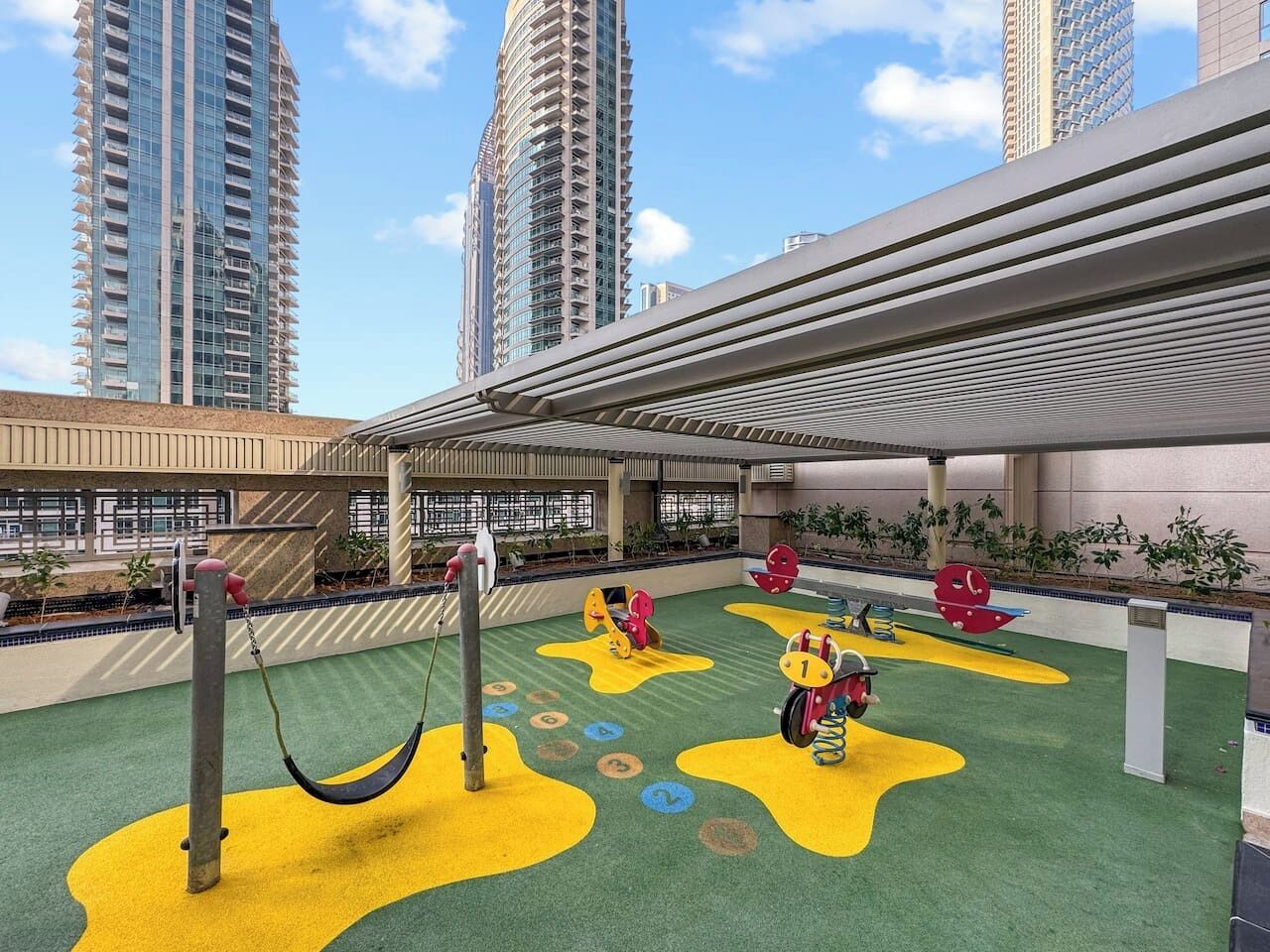 An outdoor play area with swings and climbing structures, surrounded by a building with a modern design and high-rise buildings in the background.