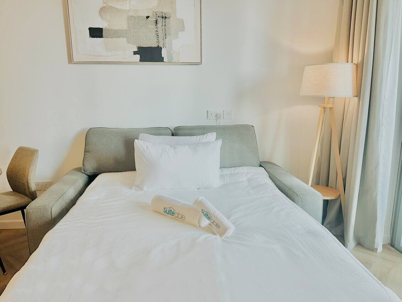 A neatly made bed with white linens and a rolled towel, a grey upholstered armchair, a small wooden nightstand with a lamp, and a painting on the wall.
