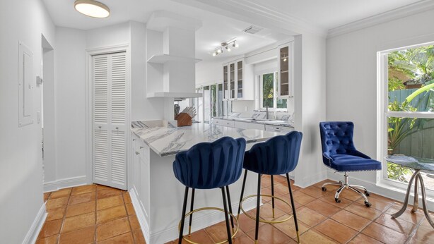 A modern kitchen with white cabinetry, a marble countertop, and a large window. There are two blue bar stools and a blue chair. - Charming 2-bedroom bungalow in lovely Delray Beach with AC (Delray Beach)