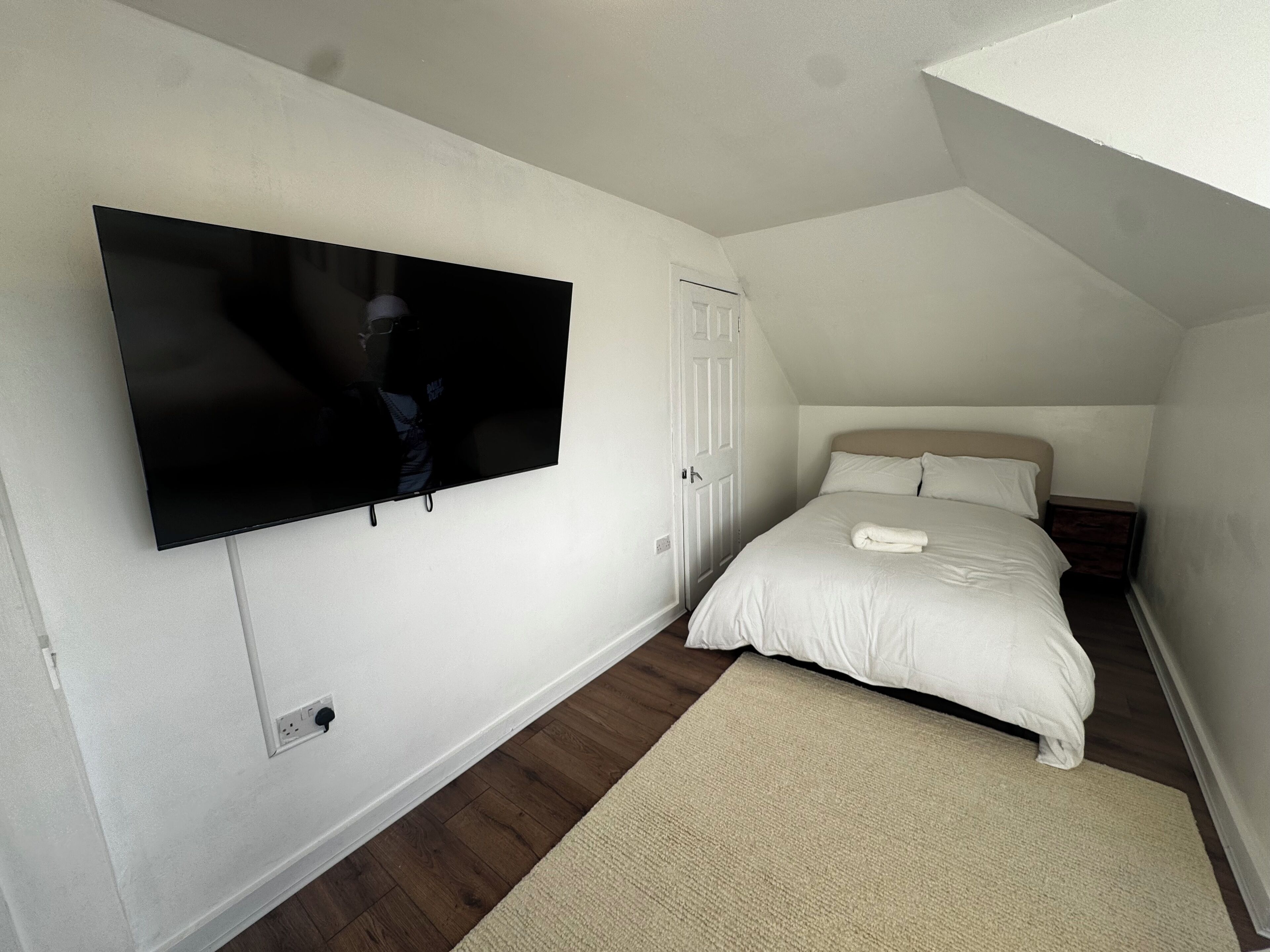 Un dormitorio con cama, televisor montado en la pared, una puerta y una mesita de noche.