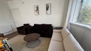 A modern living room with a beige sofa, a dark brown armchair, and a round coffee table. There are two framed artworks on the wall and a window providing a view of the outdoors. - Spacious 4BR Home Near Edinburgh Airport and City Centre – Parking (Edinburgh)