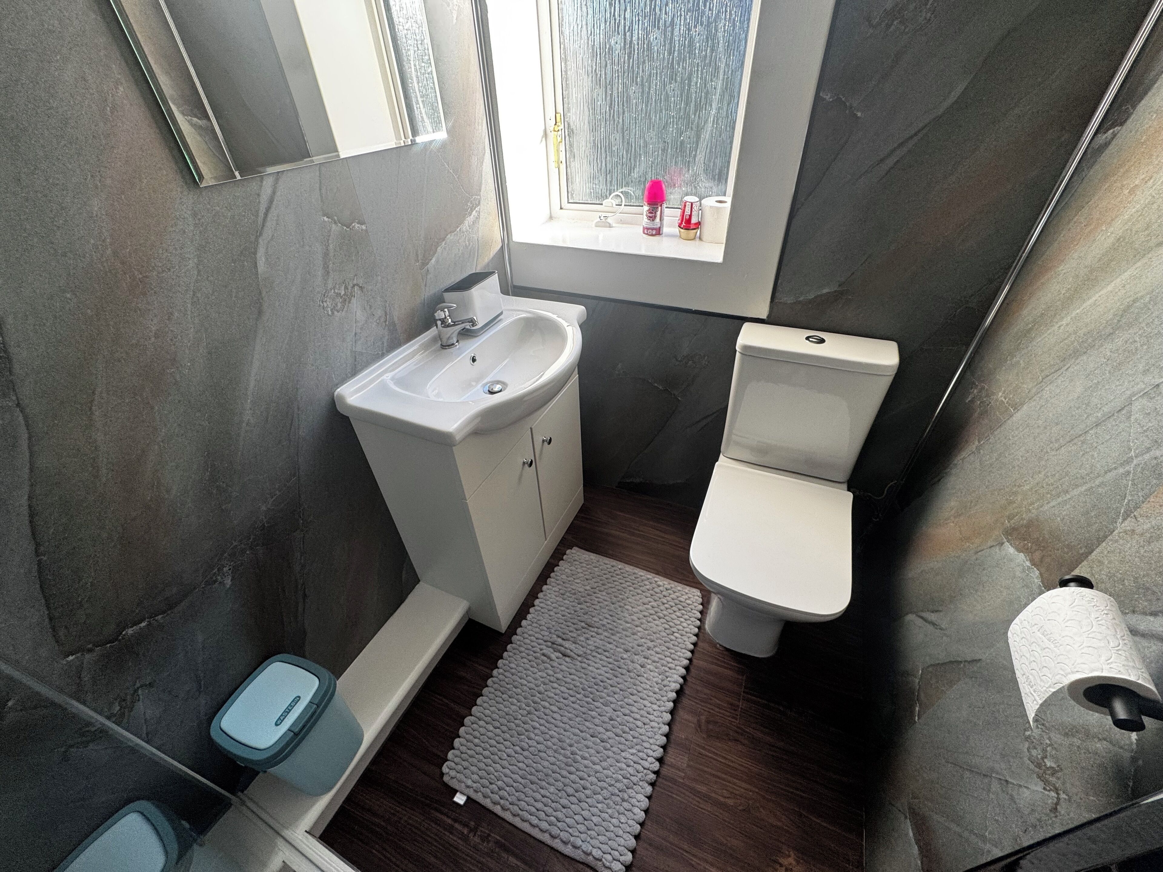 Ein kleines Badezimmer mit Waschbecken, Toilette und Dusche in Weiß. Ein kleines Fenster lässt natürliches Licht herein, und ein Handtuchhalter ist zu sehen.
