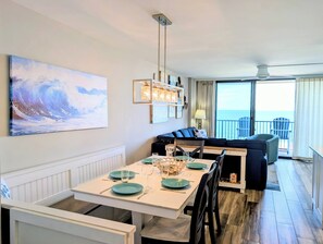 Un comedor con una mesa blanca dispuesta para cuatro personas, un sofá negro y vista al océano desde un balcón.