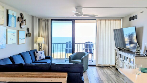 Una sala de estar con sofá, sillones, una mesa de centro, televisor y un balcón con vista al océano.