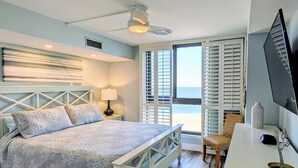 Un dormitorio con cama, escritorio, silla, lámpara, ventilador de techo y vista a la playa.