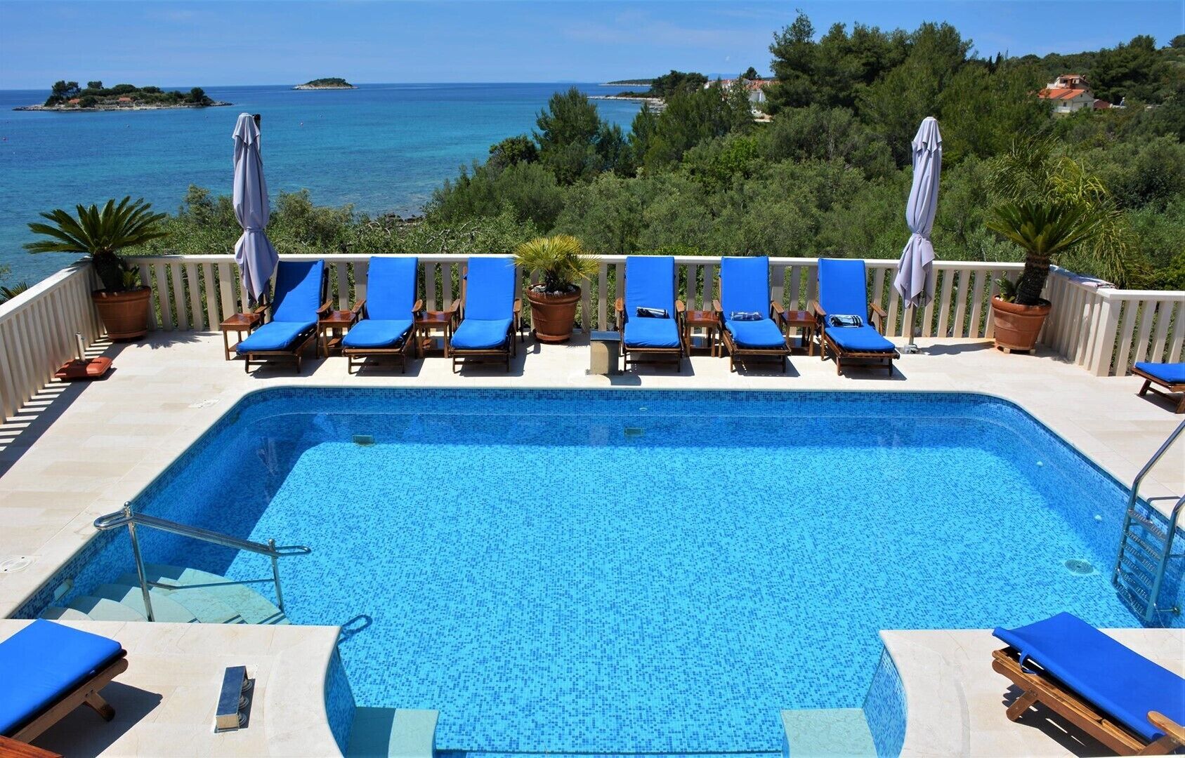 Piscina com azulejos azuis, rodeada por espreguiçadeiras e guarda-sóis, com vista para uma área costeira com árvores e casas.