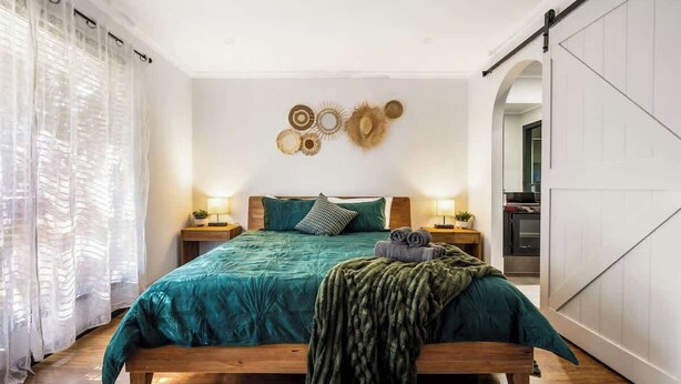 Quarto com cama de madeira, roupa de cama verde-água, duas luminárias de cabeceira, um adorno circular na parede e uma porta deslizante à direita.