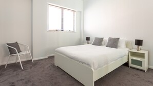 A bedroom with a white bed, a nightstand with a lamp, a chair, and a window with blinds. - Chic Unit With Rooftop Terrace Near Sights & Shops (Sydney)