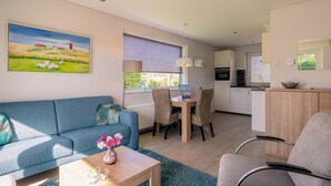 Un salon moderne avec un canapé, une table basse et un tableau représentant un paysage. On y trouve également un coin repas avec des chaises et une table, ainsi qu’une cuisine à l’arrière-plan.