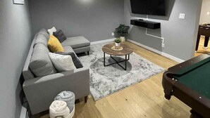 Un salon moderne avec un canapé gris, une table basse, un billard et un téléviseur à écran plat fixé au mur.