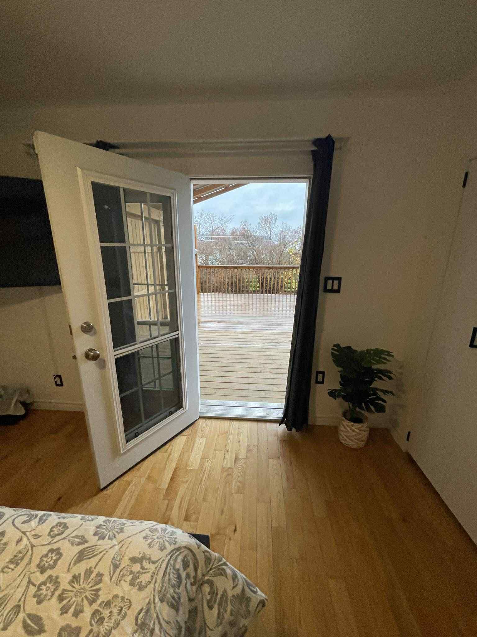 Une pièce avec un plancher en bois, un lit recouvert d’une couvre-lit à motifs floraux, une porte-fenêtre coulissante donnant sur une terrasse extérieure et une plante en pot.