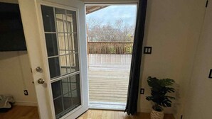 Une pièce avec un plancher en bois, un lit recouvert d’une couvre-lit à motifs floraux, une porte-fenêtre coulissante donnant sur une terrasse extérieure et une plante en pot.
