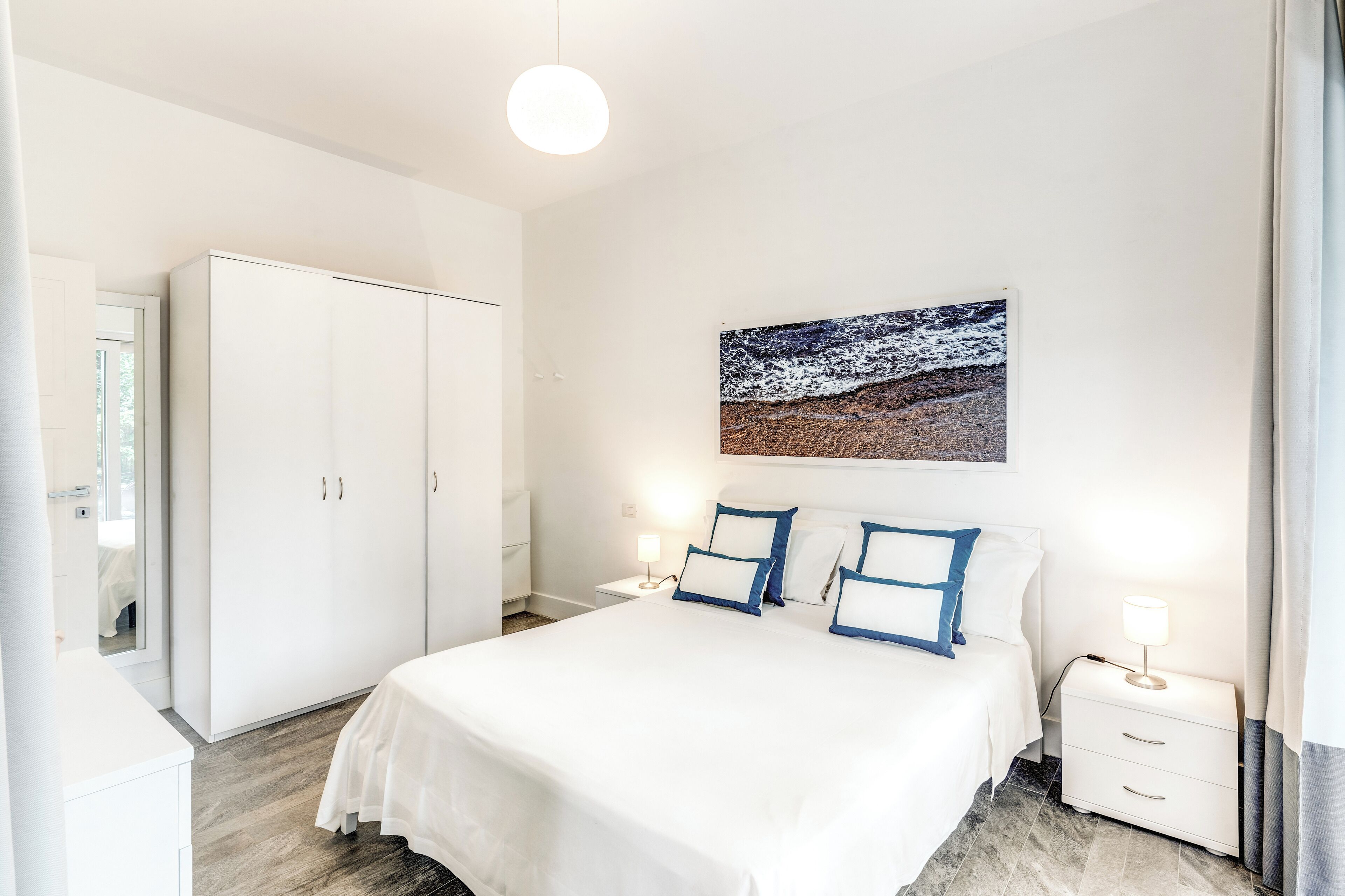 A modern bedroom with a large bed, white wardrobe, bedside tables, and a wall-mounted artwork.