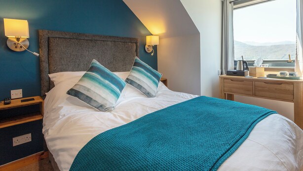 A neatly made bed with a teal throw blanket, two pillows, and a wooden nightstand with a lamp. - Lochbroom Lodge (Ullapool)