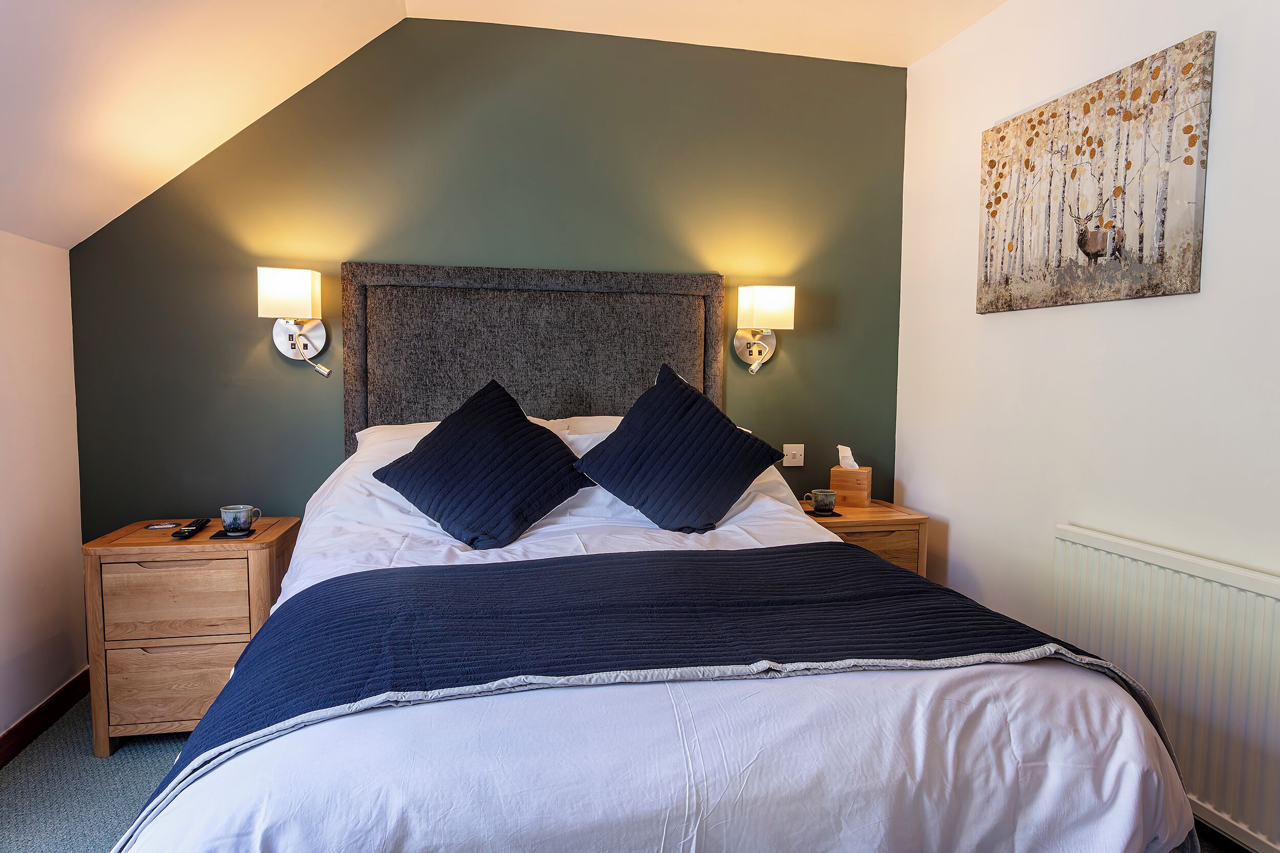 Ein ordentlich gemachtes Bett mit dunkelblauer Bettdecke und zwei Kissen, flankiert von Nachttischen mit Lampen und einer Tasse.