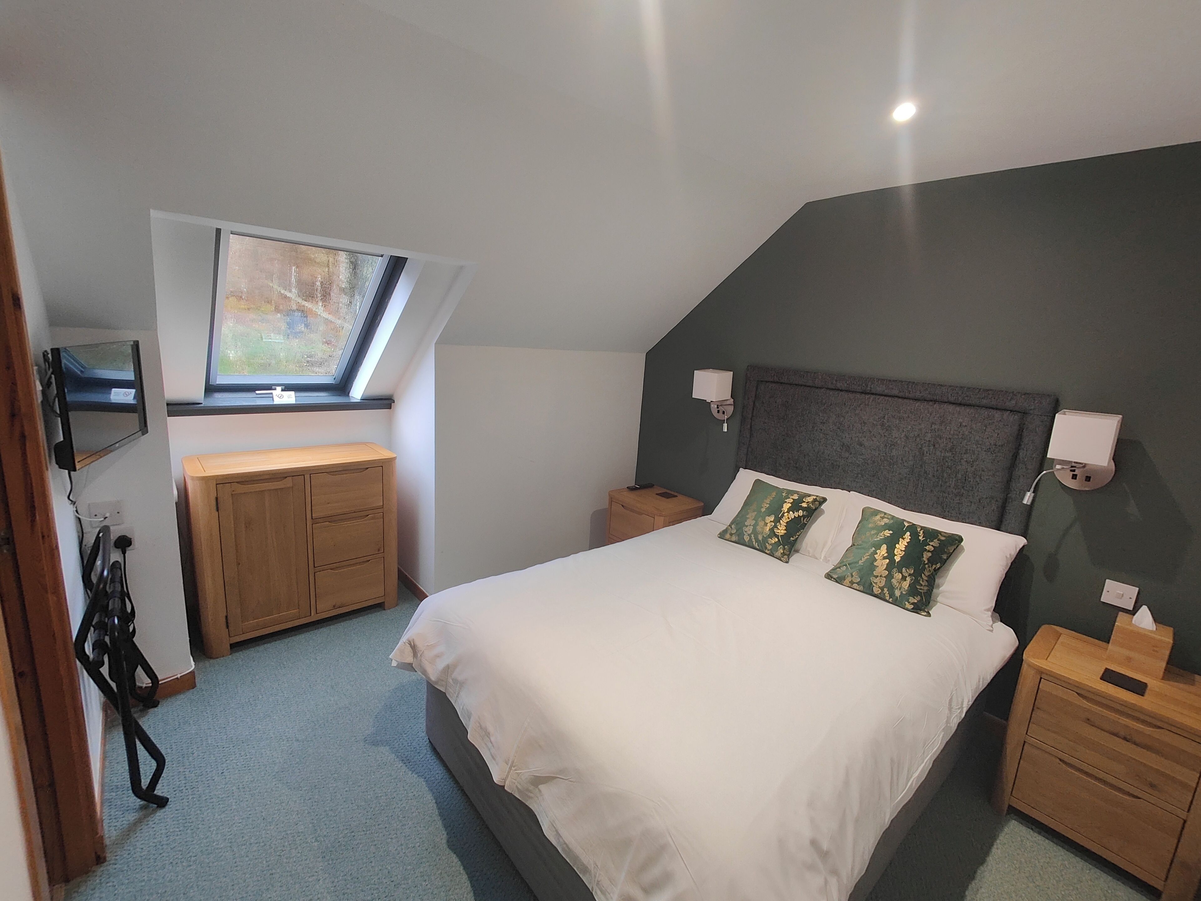 Un dormitorio con cama, mesitas de noche, escritorio con silla, ventana y televisor montado en la pared.