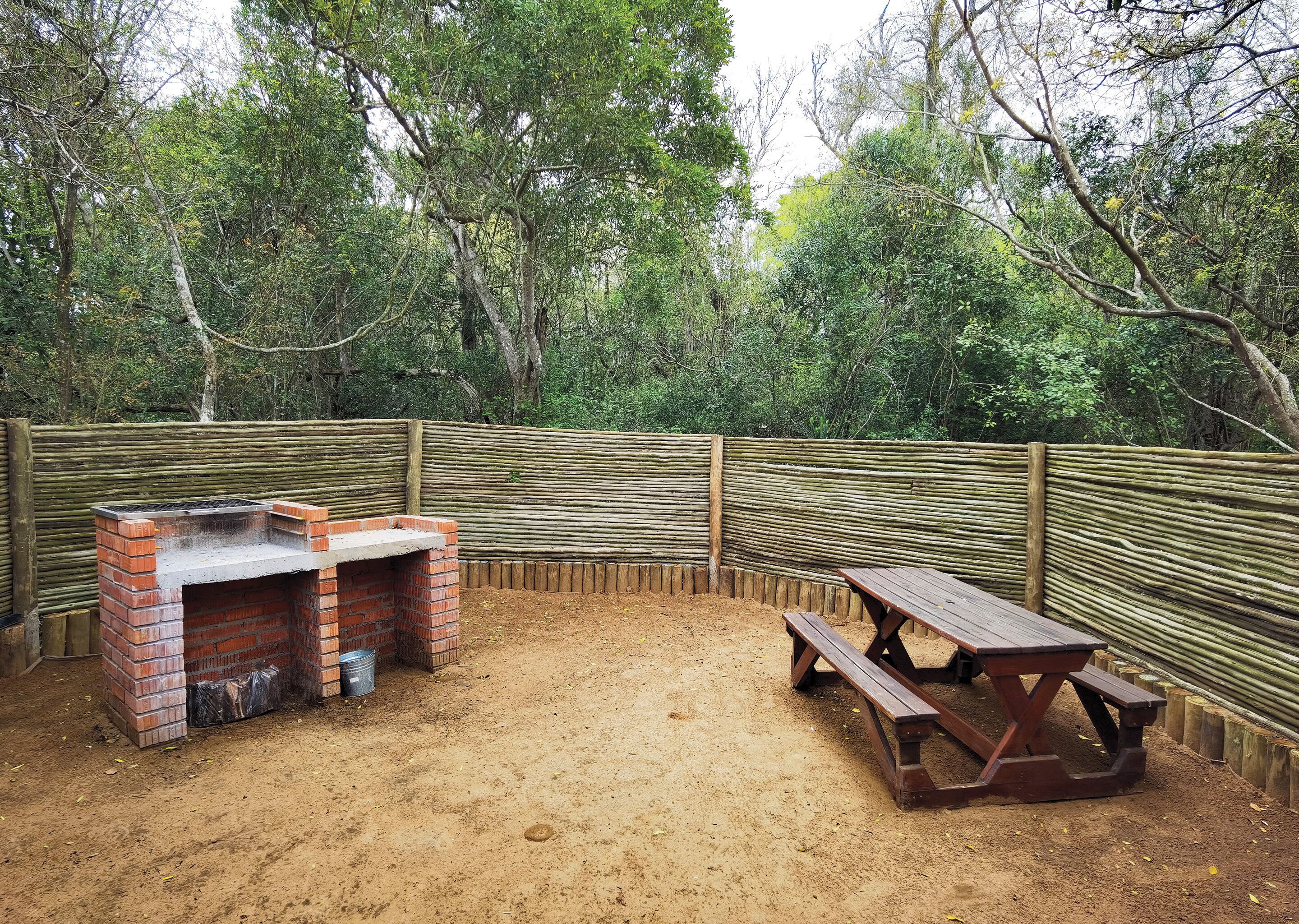 Un espace barbecue extérieur avec un gril en briques et une table de pique-nique en bois, entouré d’une clôture en bois et de nombreux arbres.