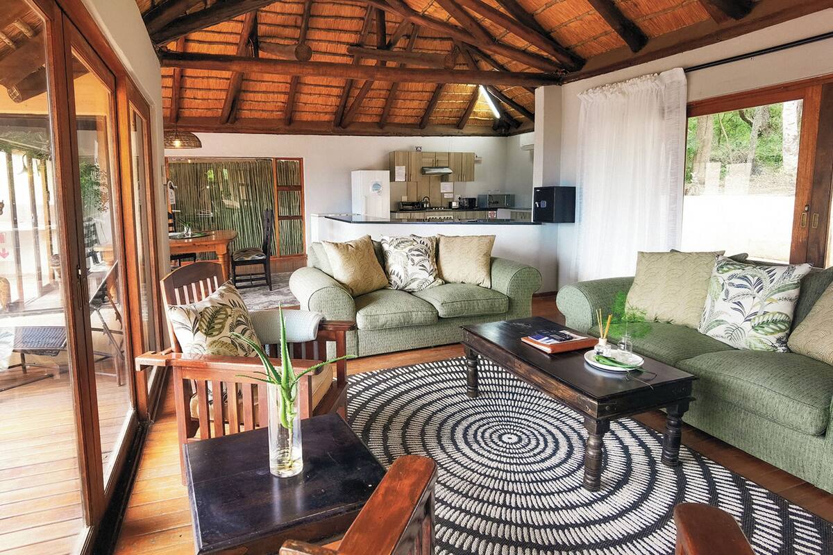 Uma sala de estar acolhedora com um sofá verde, mesa de centro de madeira e um tapete estampado. Uma área de jantar com mesa e cadeiras de madeira é visível ao fundo.