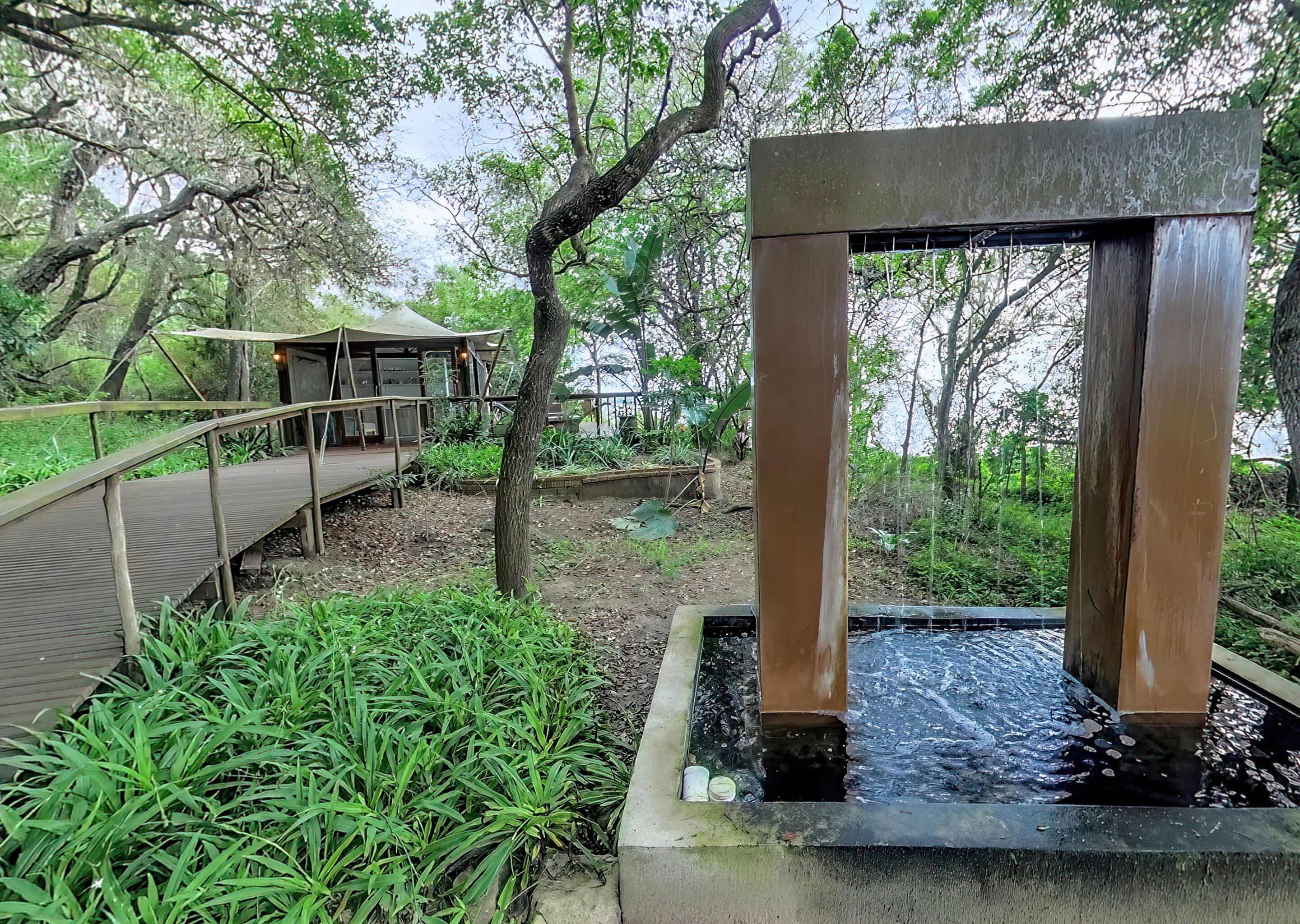 A wooden bridge over a water feature with a metal railing, surrounded by greenery.