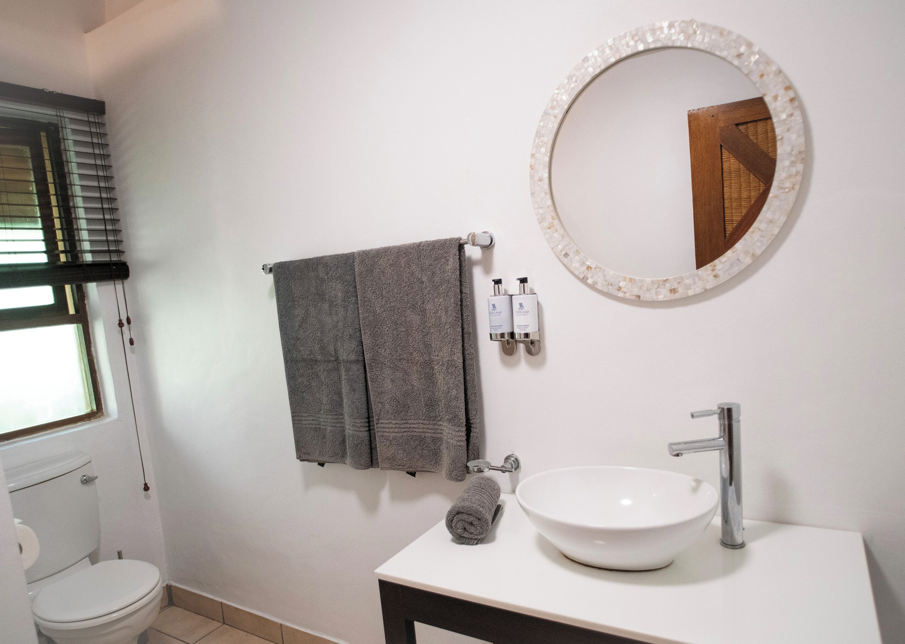 Une salle de bain avec un lavabo blanc, un miroir rond orné d’un cadre en mosaïque et deux serviettes suspendues à un porte-serviettes.