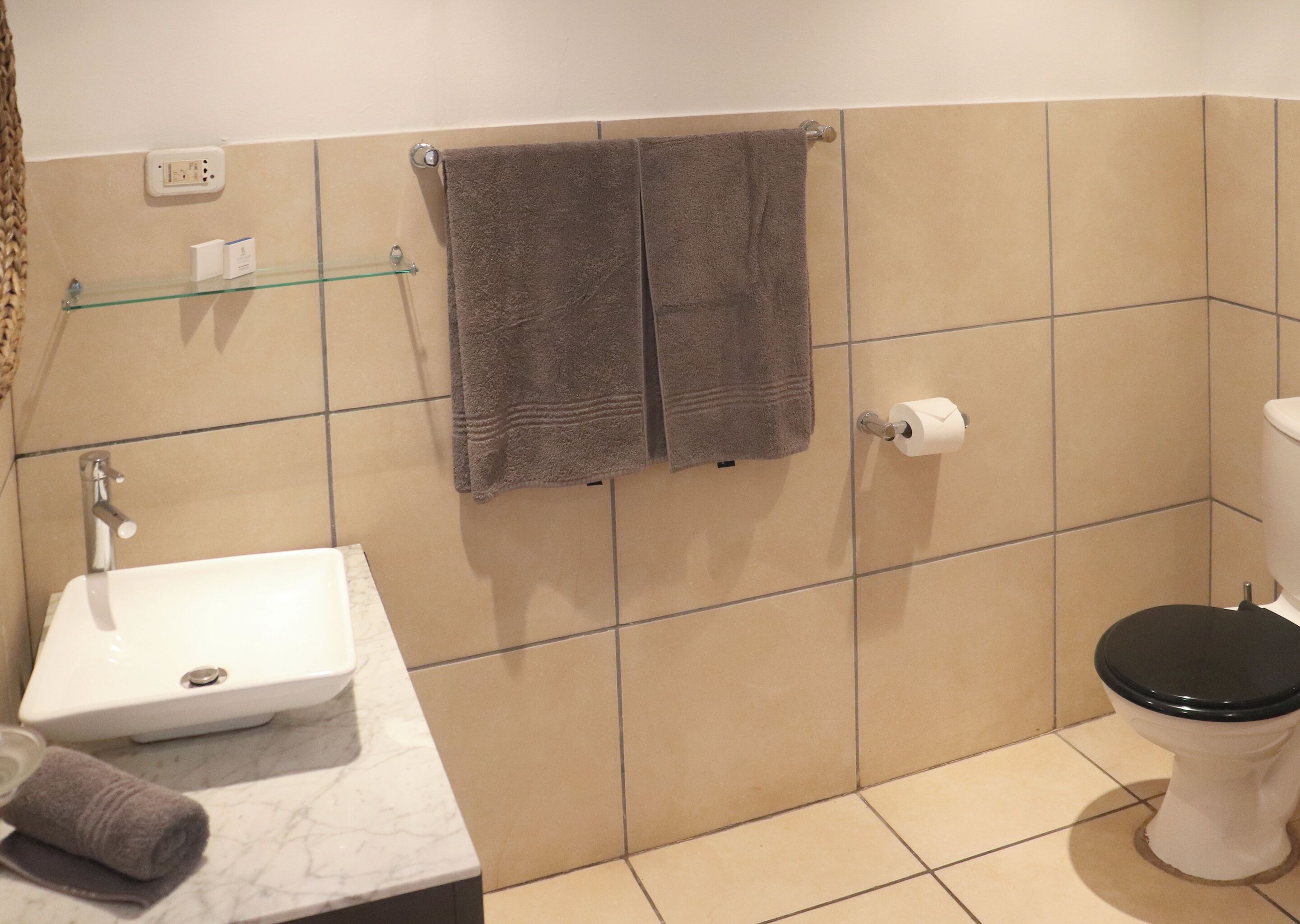 Une salle de bain avec un lavabo blanc, deux serviettes, une étagère en verre, des toilettes et des murs recouverts de carreaux beiges.