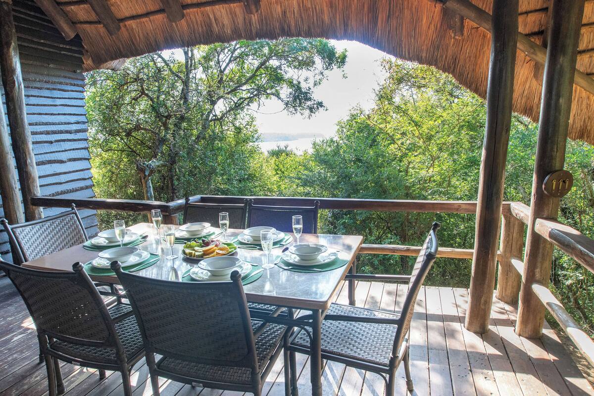 Um deck de madeira com mesa posta para refeições, rodeado por cadeiras, com vista para uma paisagem verde exuberante.