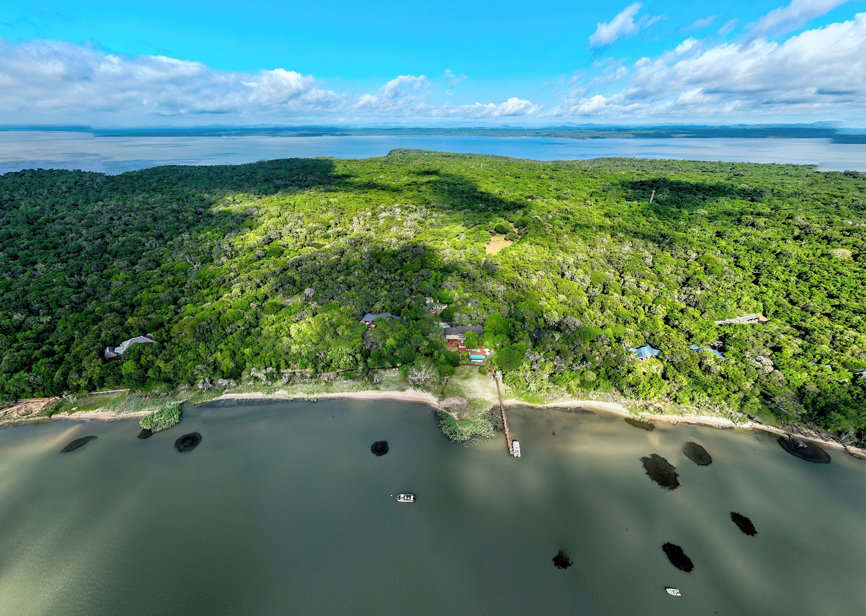 An aerial view of a coastal area with dense greenery, a beach, and a small body of water with boats.