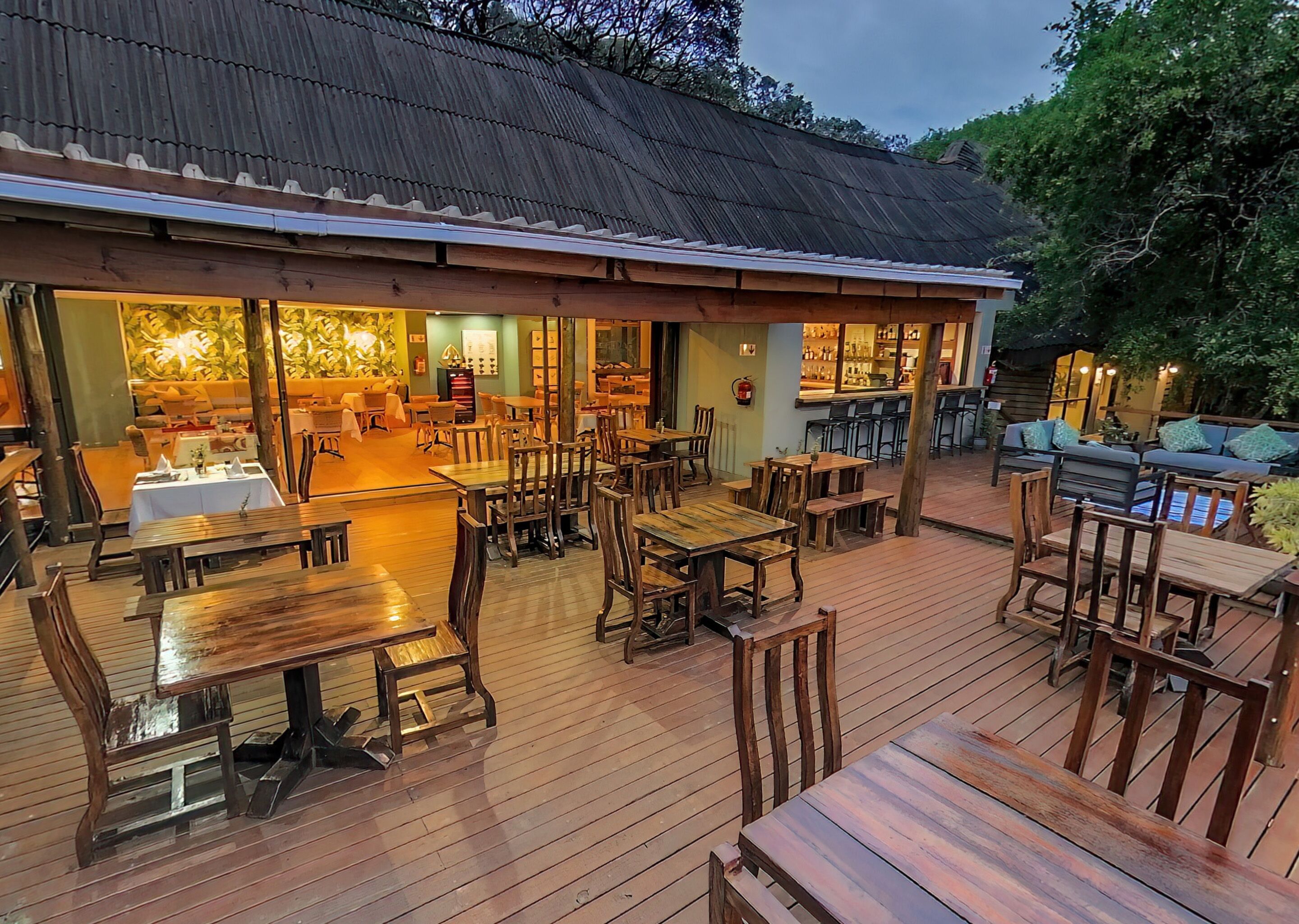 An outdoor dining area with wooden tables and chairs, a bar area with stools, and a covered seating area with cushions.