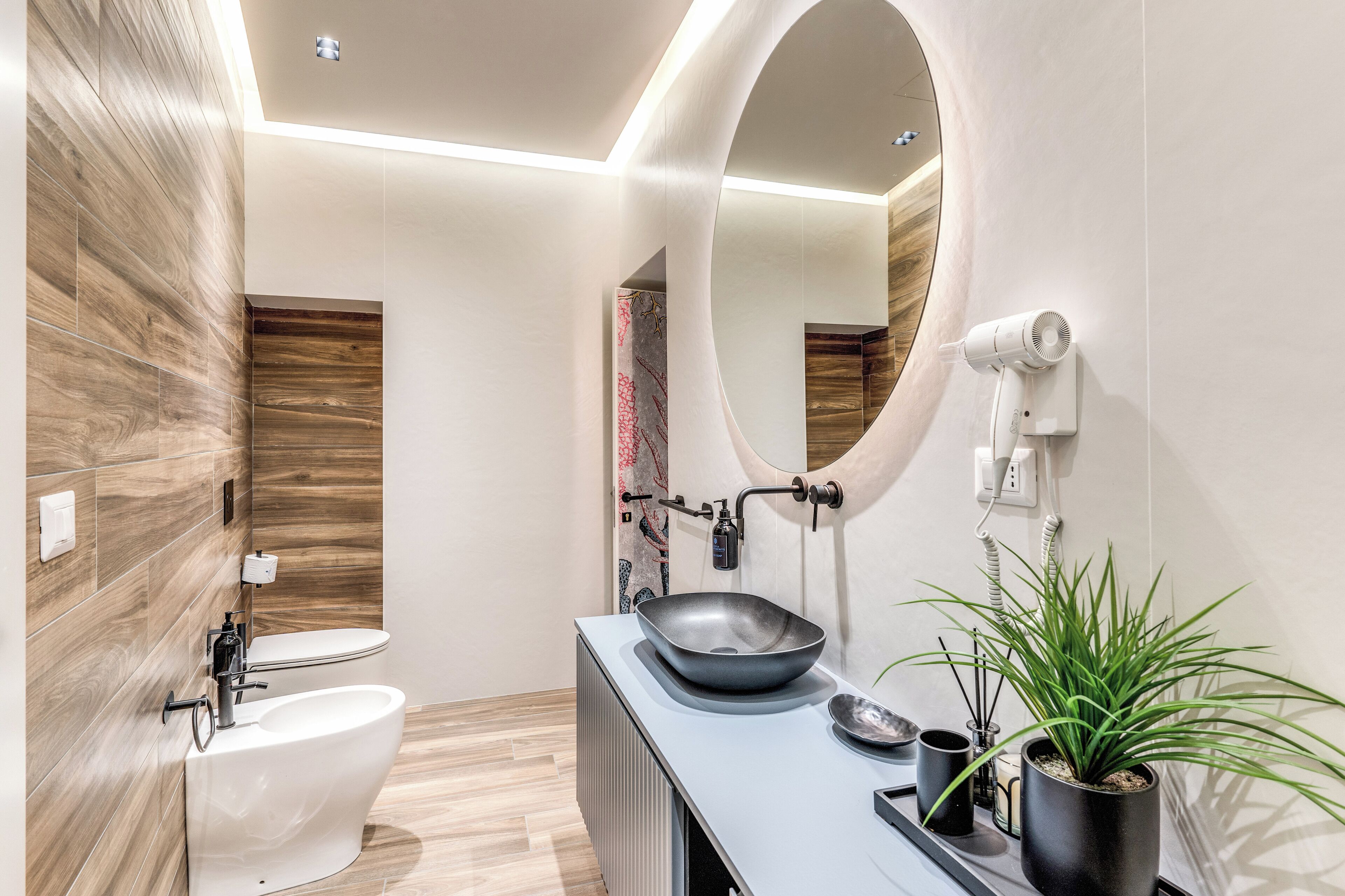 Un baño moderno con pared de madera, un inodoro blanco, un lavabo negro con recipiente, un espejo y una planta en maceta.