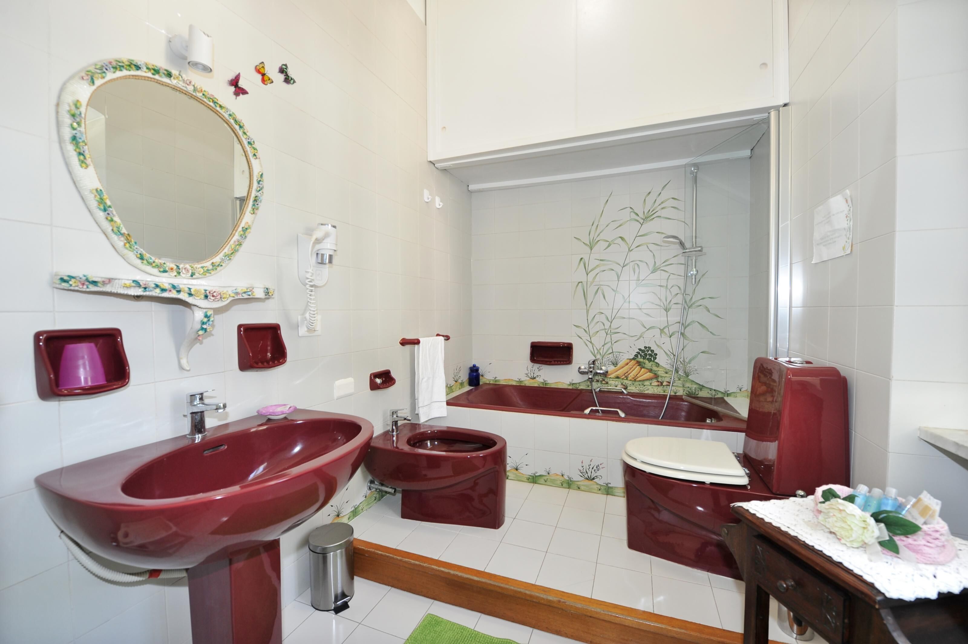 A bathroom with a red sink, a toilet, and a bathtub. There is a mirror with a floral frame, a towel rack, and a small trash can.