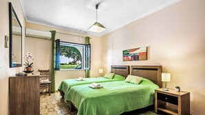 Une chambre d’hôtel avec deux lits, un bureau, une chaise, une petite table, une table de chevet, une lampe, une plante en pot, une fenêtre avec vue et un tableau au mur.