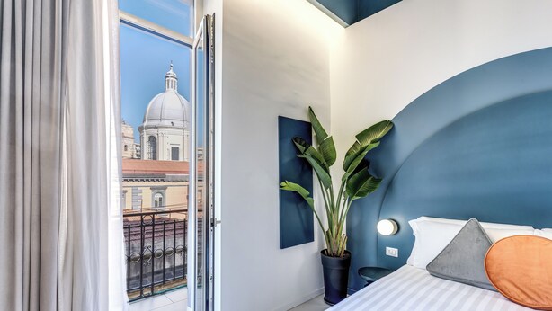 Una habitación de hotel moderna con una cama, una planta en maceta y vistas de un paisaje urbano a través de una puerta abierta.