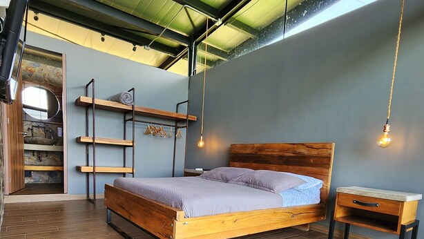 A bedroom with a wooden bed, a nightstand, a shelf with hanging clothes, and a mirror. - Landscape Suite (Cerritos)