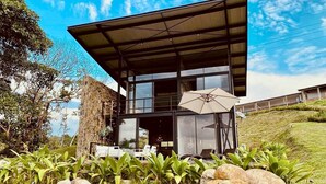 A modern building with a glass facade, a covered patio with outdoor furniture, and a stone wall. - Landscape Suite (Cerritos)