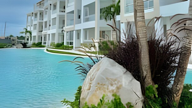 Et moderne hotel med flere balkoner med udsigt over en swimmingpool, omgivet af frodig vegetation og palmer.