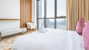 Ein Hotelzimmer mit einem großen Bett, einem Sessel, einer Bank und Blick auf eine Stadtkulisse durch ein großes Fenster.