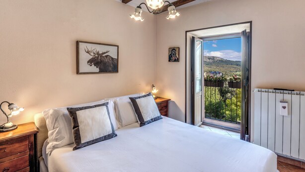 Ein Schlafzimmer mit Bett, Nachttischlampen, einem Bild von einem Elch im Bilderrahmen, einer kleinen religiösen Ikone und Blick auf einen Balkon mit Geländer und Bepflanzung.