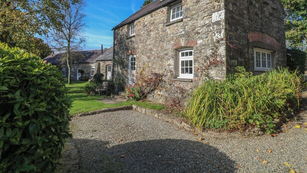 A stone house with a gravel driveway, surrounded by greenery and a clear blue sky. - Carthouse Cottage (Clarbeston Road)